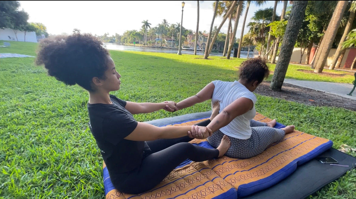 Load video: Preview of traditional Thai  massage therapy session outside at the park. Performed on traditional thai mat fully clothed. Guided deep breathing and passive stretching moving through a series of different yoga poses.